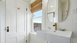 A bathroom with a white sink, a mirror, and a window with a view of buildings. - Boothbay Harbor King Suite - Room 4 at Greenleaf Inn - Fireplace, Harbor Views! (Boothbay Harbor)
