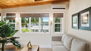 A room with a wooden ceiling, large windows with blinds, a beige sofa, a wooden coffee table, and a potted plant. - Boothbay Harbor King Suite - Room 4 at Greenleaf Inn - Fireplace, Harbor Views! (Boothbay Harbor)