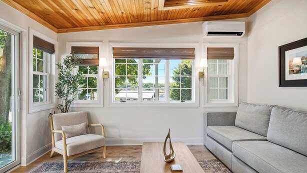 A room with a wooden ceiling, large windows with blinds, a grey sofa, a beige armchair, and a patterned rug. - Boothbay Harbor King Suite - Room 4 at Greenleaf Inn - Fireplace, Harbor Views! (Boothbay Harbor)