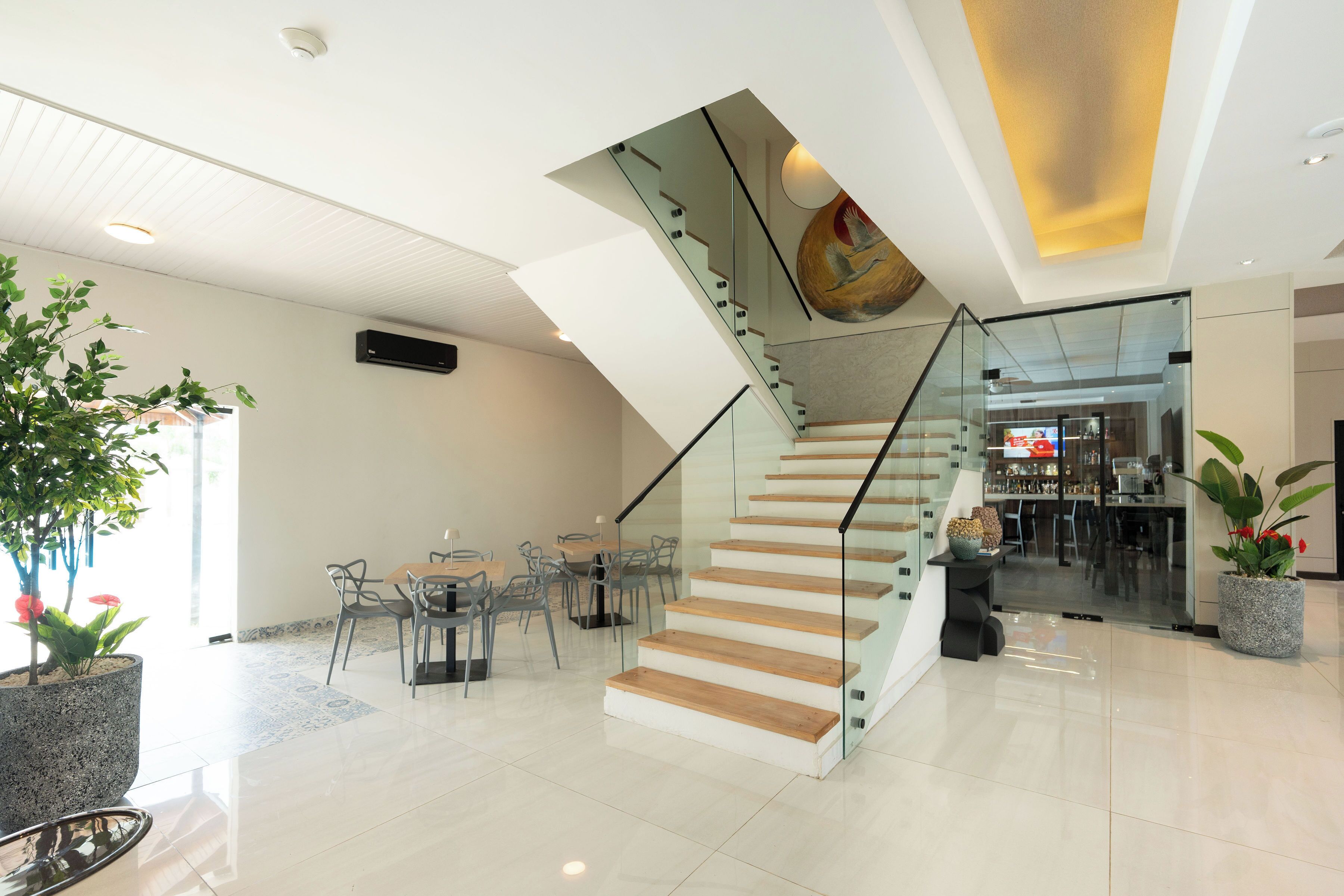 Un moderno vestíbulo de hotel con una escalera de cristal, un comedor con mesas y sillas, y plantas en macetas.