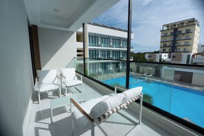 Un balcon doté d’une balustrade en verre donnant sur une piscine, avec des chaises longues blanches et un bâtiment moderne en arrière-plan.