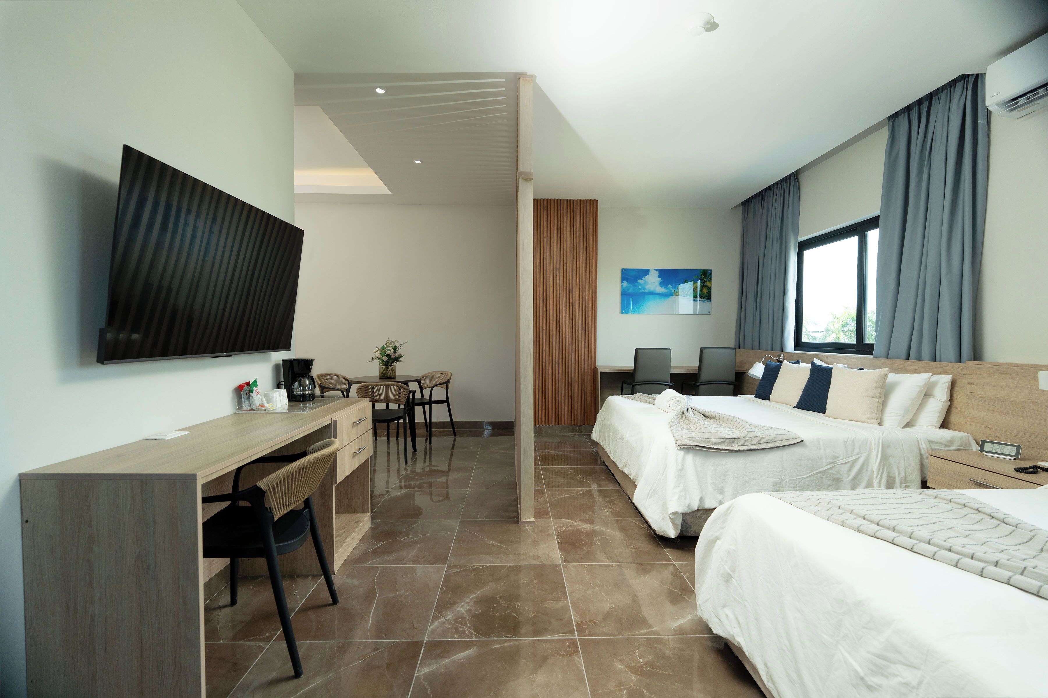 A modern hotel room with a flat-screen TV, a desk with a chair, two beds, a wooden headboard, and a large window with curtains.