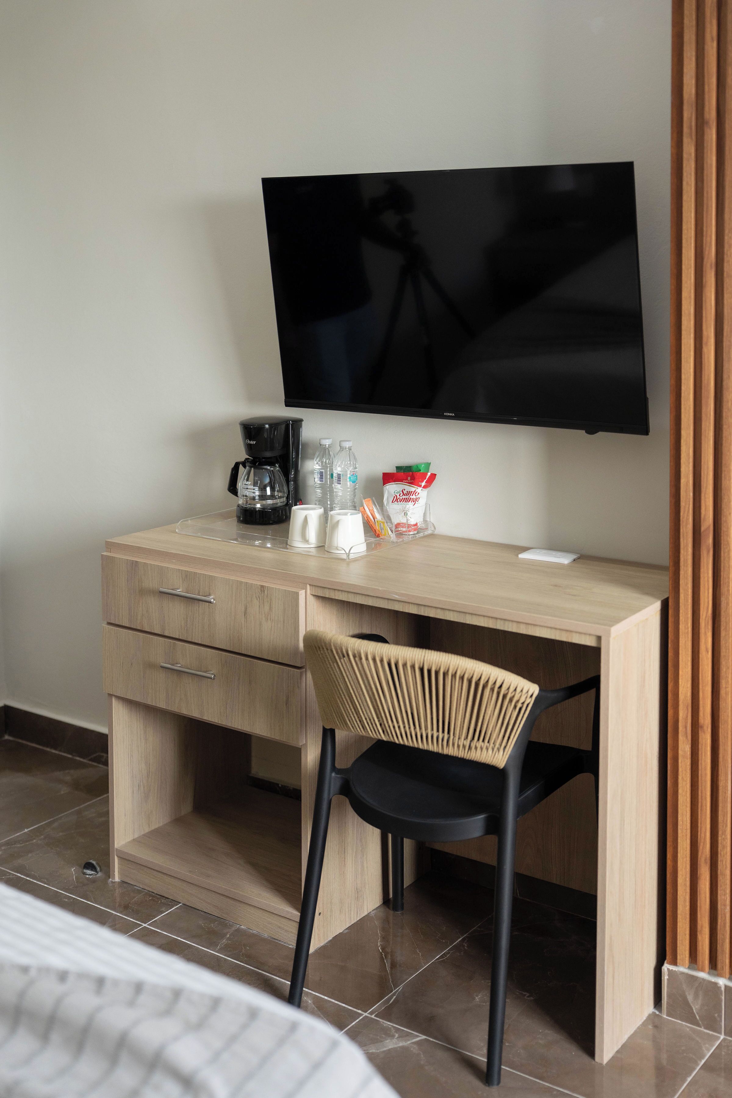 A hotel room with a wooden desk, a chair, a television mounted on the wall, a coffee maker, and some snacks.