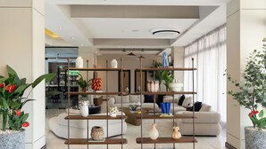Uma sala de estar moderna com um sofá seccional, uma mesa de centro e prateleiras decorativas. Há plantas em vasos e uma grande janela que proporciona iluminação natural.