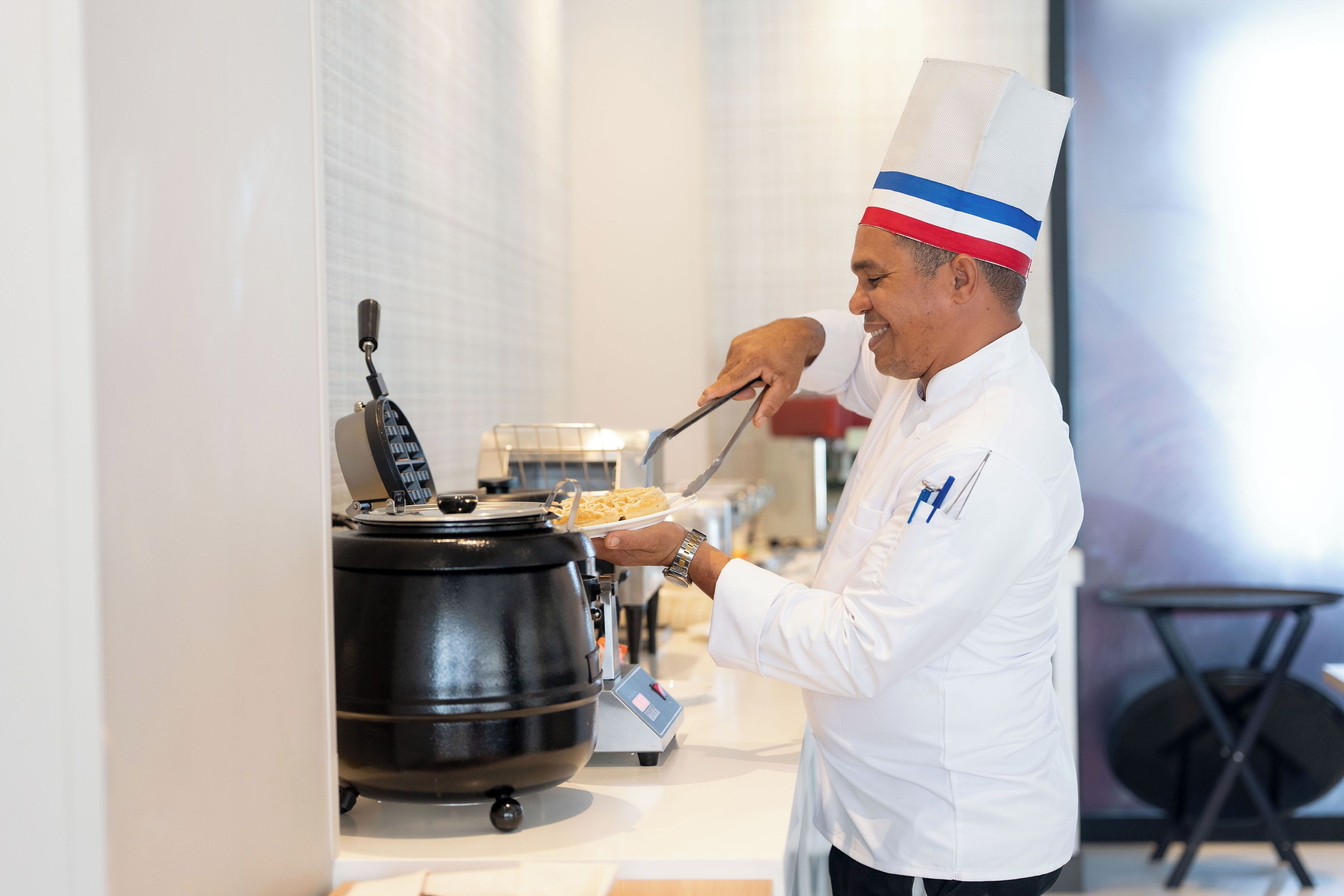 Ein Koch in weißer Uniform und Kochmütze serviert Essen aus einem Topf.