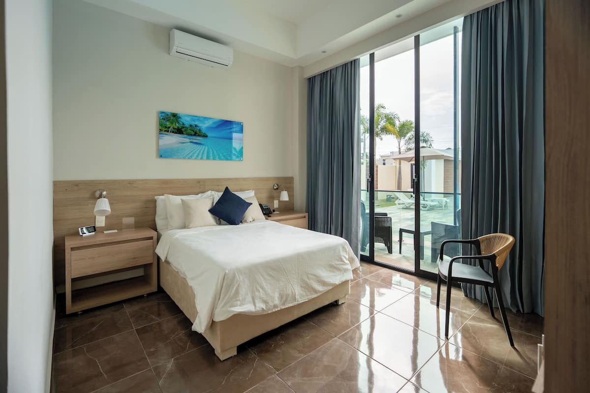 A hotel room with a large bed, a bedside table with a lamp, a chair, and a view of the outdoors through a sliding glass door.