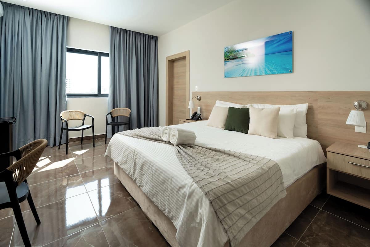 A hotel room with a large bed, a desk with a lamp, two chairs, and a window with curtains.