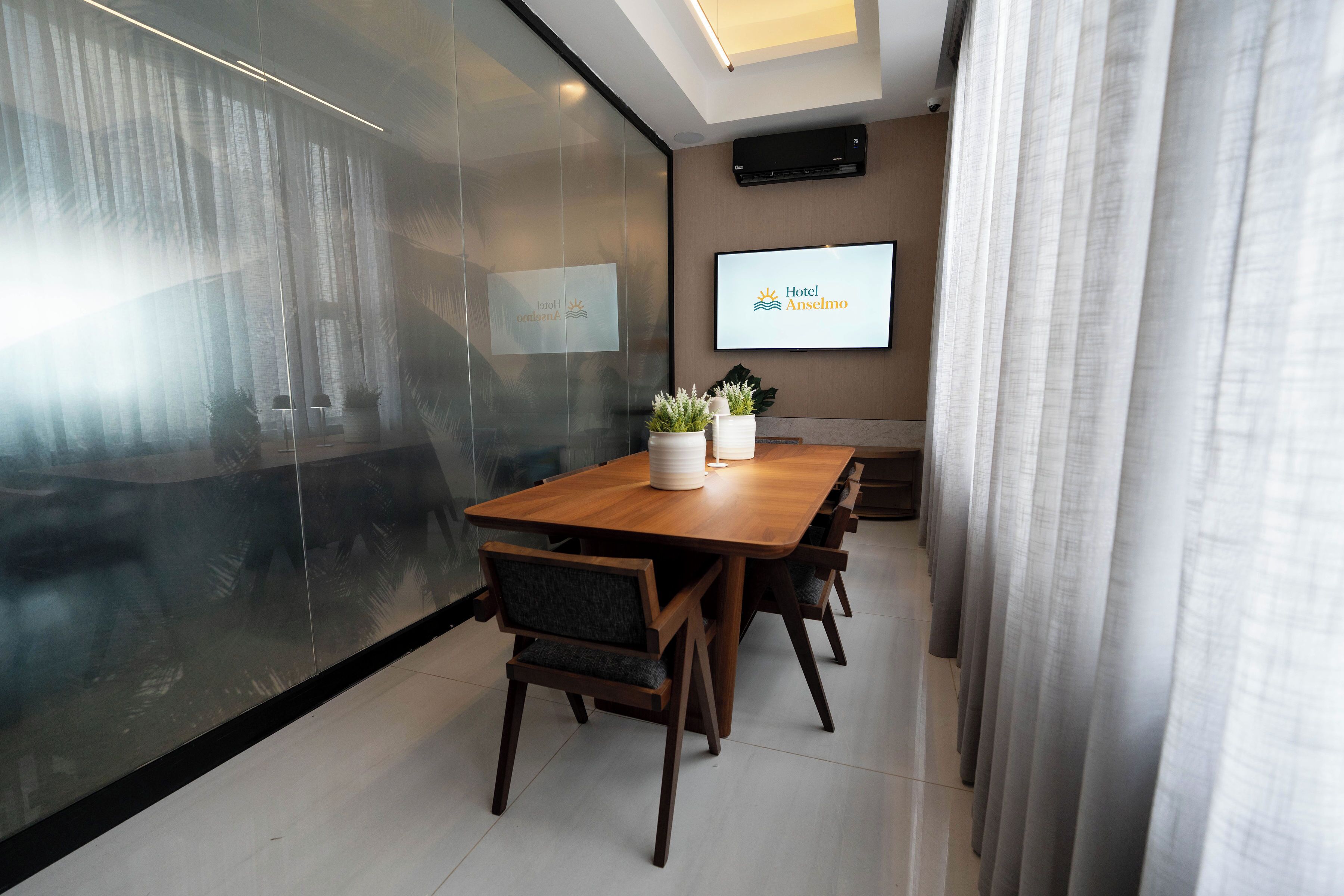 A modern meeting room with a large table, chairs, a TV screen displaying 'Hotel Anselmo', and a glass wall with a view.