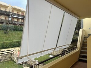 Un balcon doté d’un store blanc rétractable, d’une rambarde en métal et offrant une vue sur un jardin bien entretenu et sur des bâtiments en arrière-plan.