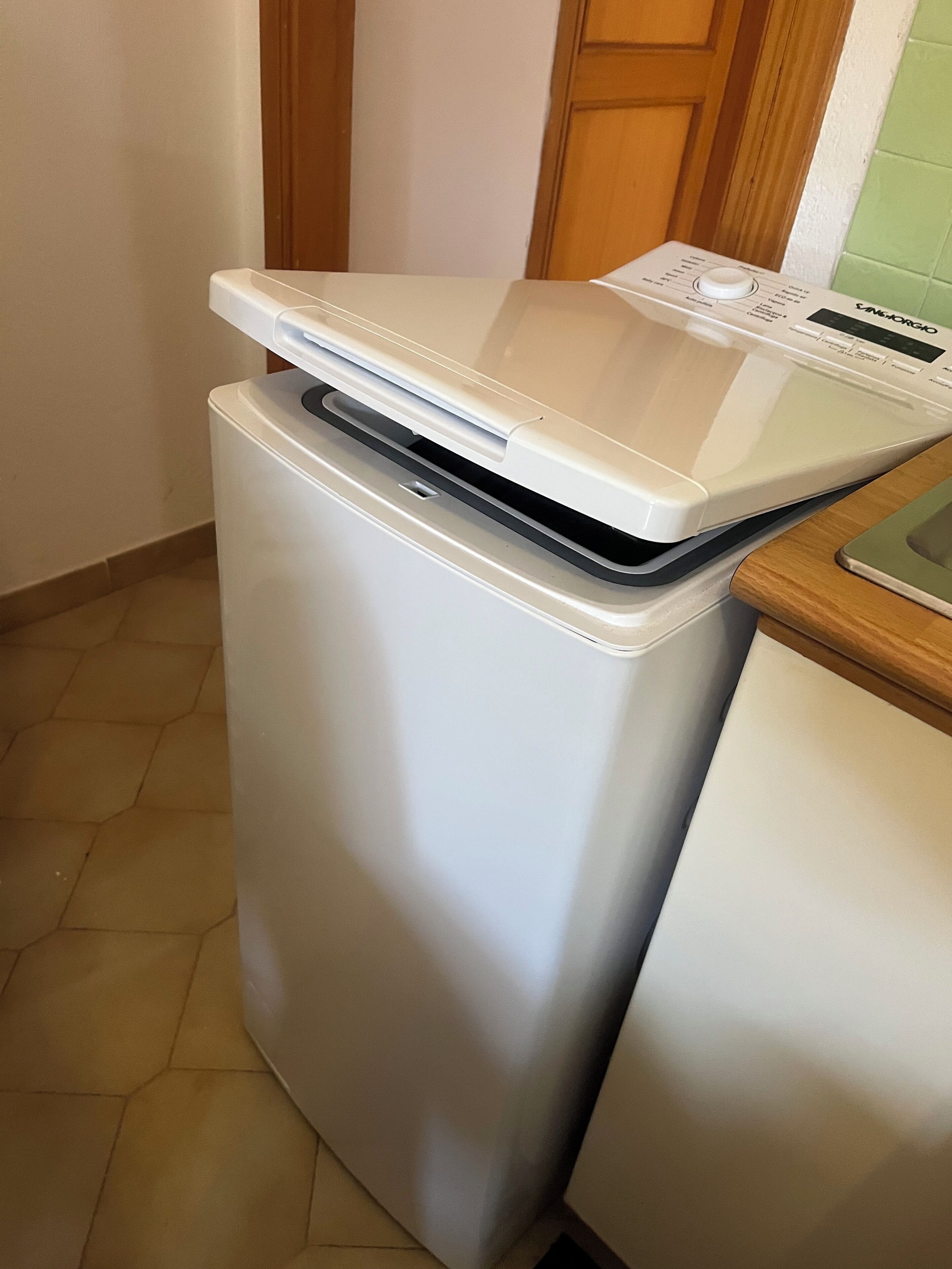A compact, white, front-loading washing machine with a control panel on top, situated in a kitchen with a tiled floor and wooden cabinets.