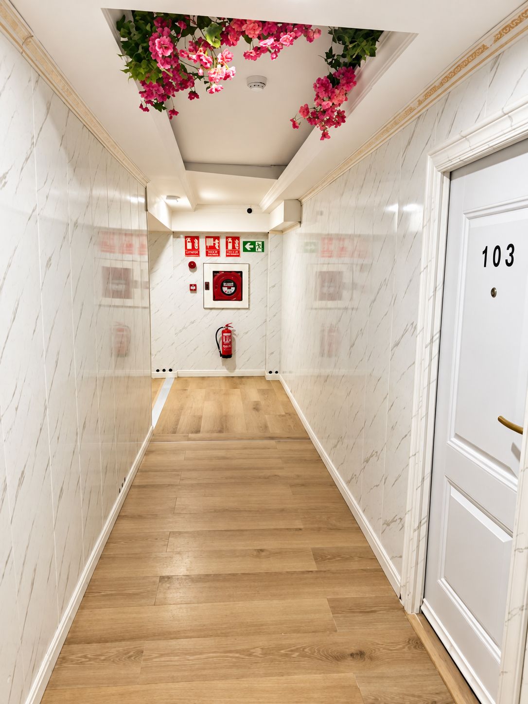 Un pasillo con piso de madera, paredes blancas con diseños tipo mármol, un extintor y una puerta con la etiqueta '103'.
