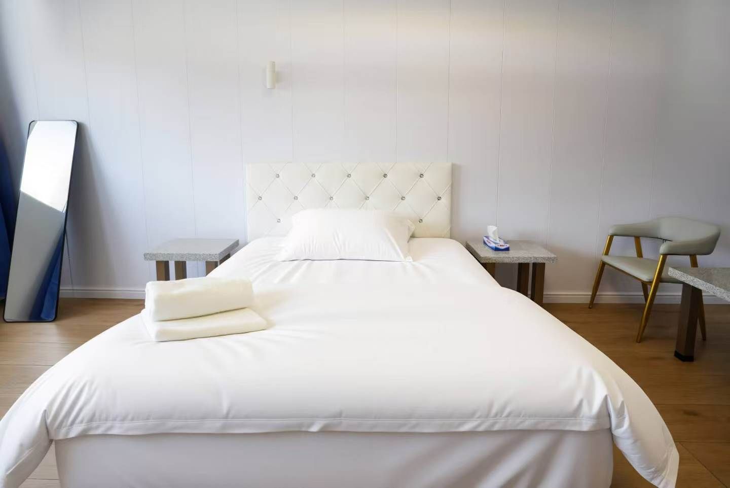 A neatly made bed with pillows, a bedside table with a lamp, a chair, and a large mirror on the wall.