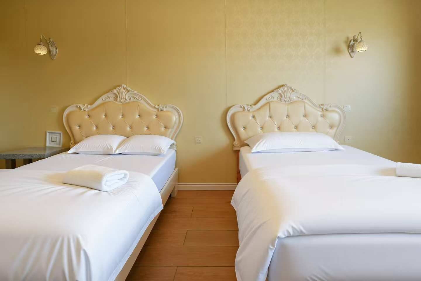 Two twin beds with white linens, a wooden floor, and a plain wall with two wall-mounted light fixtures.