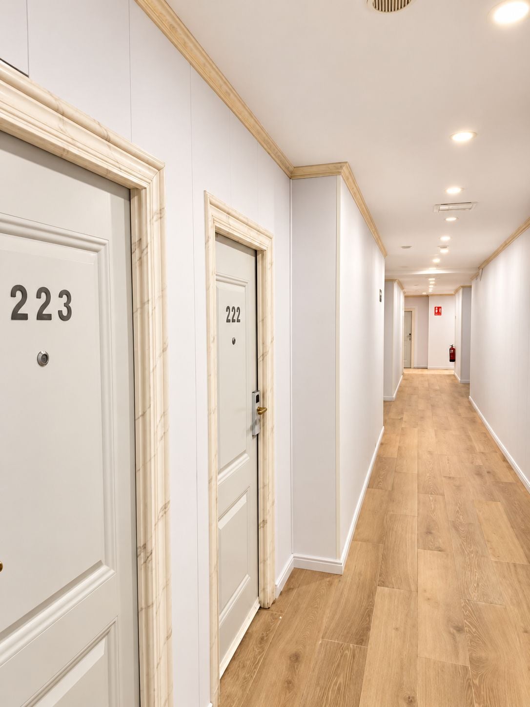 Un pasillo con puertas numeradas 223 y 222, piso de madera y luces empotradas en el techo.
