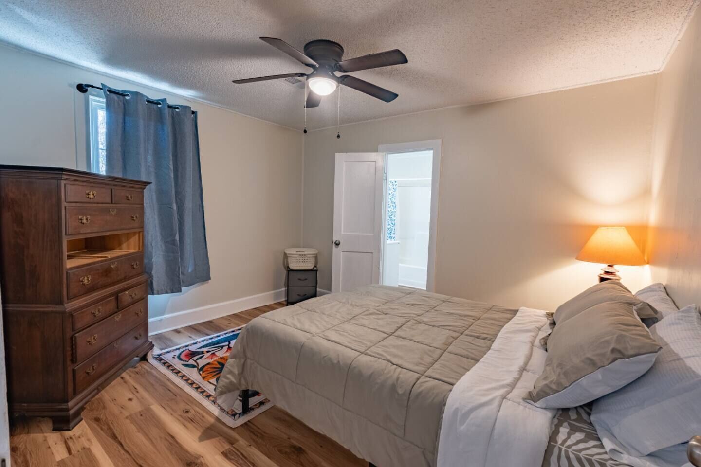 A bedroom with a bed, a dresser, a ceiling fan, a nightstand with a lamp, and a window with curtains.