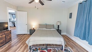 Un dormitorio con cama, dos mesitas de noche con lámparas, ventilador de techo, suelo de madera y alfombra colorida.