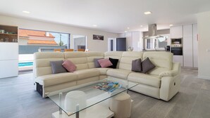 Una sala de estar moderna con un sofá seccional color beige, una mesa de centro de vidrio y una cocina con electrodomésticos integrados.