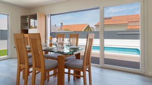 Un comedor con mesa de vidrio y sillas de madera, un aparador de vidrio y vistas a una piscina y casas.