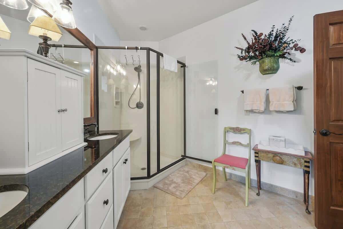 A bathroom with a glass-enclosed shower, white cabinets, a vanity with a sink, and a small table with a chair.