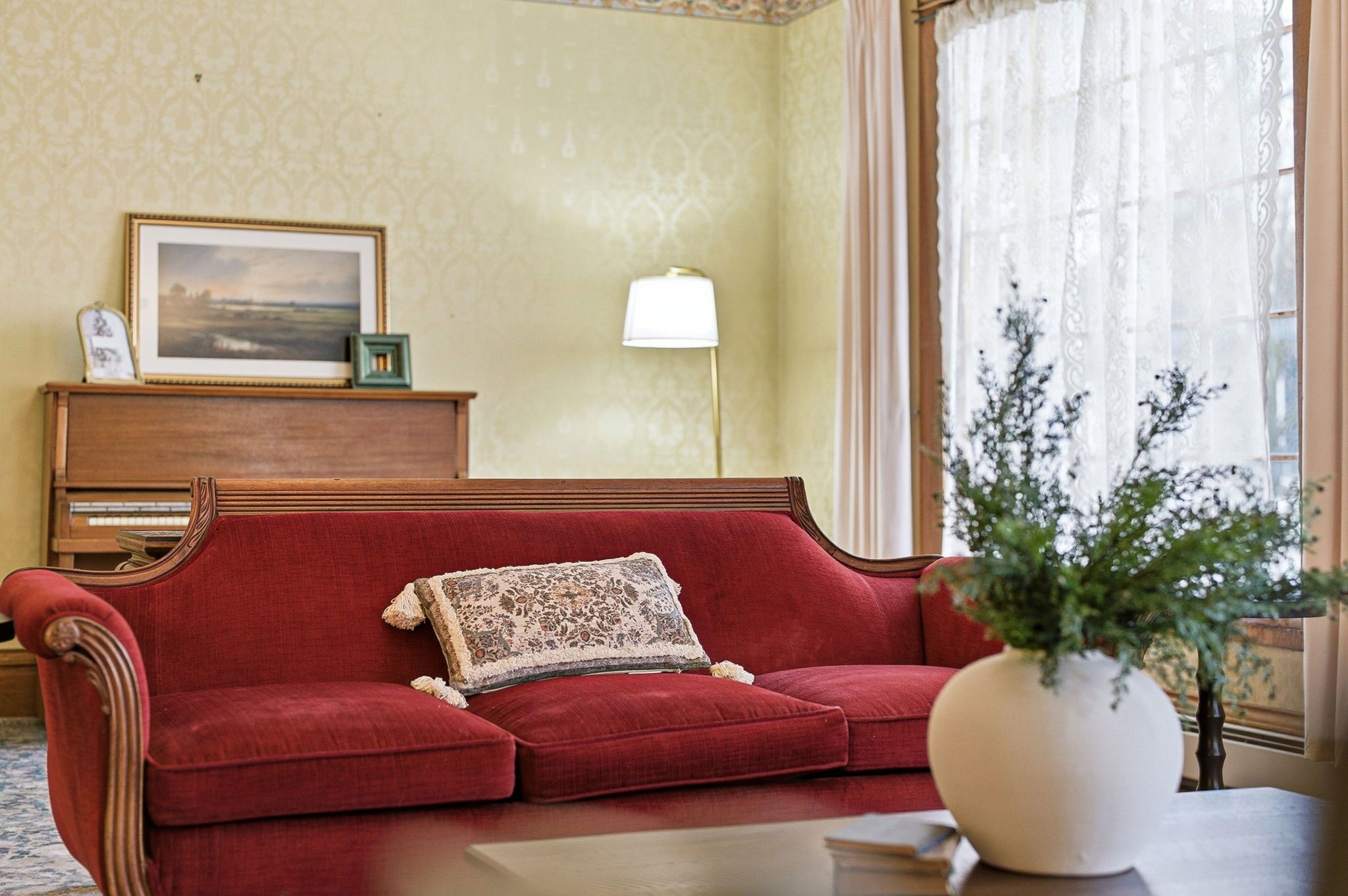 A red sofa with a patterned throw pillow, a wooden side table, a vase with green foliage, a framed picture, and a lamp on a wall.