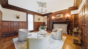A traditional room with wooden paneled walls, a fireplace, and a chandelier. There are two armchairs and a small table with a plant.