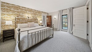 A bedroom with a large bed, a nightstand, a wall with a patterned wallpaper, a window with curtains, and a door.