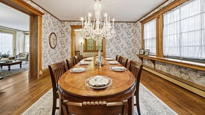A dining room with a wooden table set for six, surrounded by chairs. A chandelier hangs from the ceiling, and the walls are adorned with floral wallpaper.