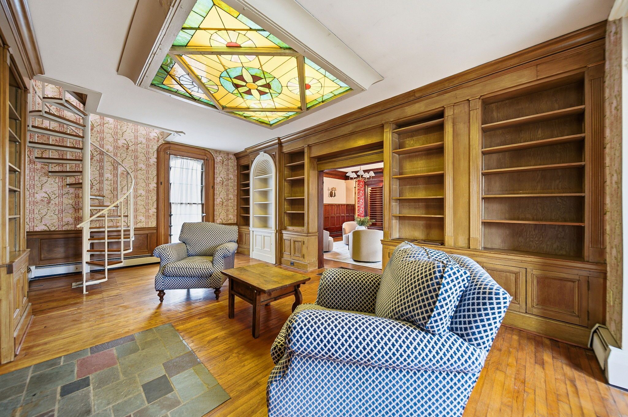 A spacious living room with a spiral staircase, built-in bookshelves, and a large stained glass window.