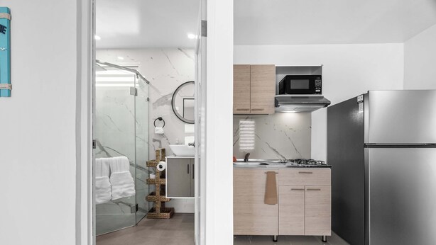 A modern kitchen with a stainless steel refrigerator, a microwave, and a gas stove. There is a glass shower enclosure with a towel rack and a wooden bath mat.