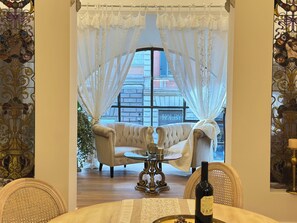 A room with a large window, a white sofa, a small round table, and a bottle of wine. - Elegant 3-bedroom apartment with a bathtub and a charming view of Rome (Roma)