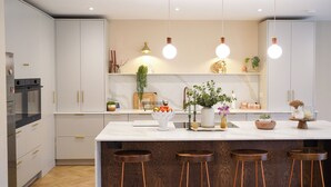 Een moderne keuken met witte kasten, een marmeren aanrechtblad en vier krukken. Er hangen hanglampen, staat een plantje en ligt een fruitschaal op het aanrecht.