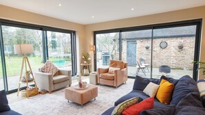 Een ruime woonkamer met grote ramen, een blauwe bank, een beige fauteuil en een klein tafeltje met decoratieve objecten.