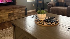 En stue med en pejs af natursten, et spisebord med en plante og fjernbetjeninger, en sofa og et vindue med persienner.