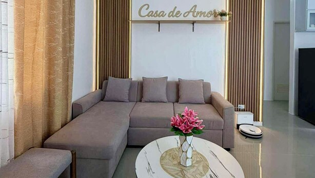 Un salon moderne avec un canapé d’angle, une table basse ronde ornée d’un vase de fleurs et un panneau mural sur lequel on peut lire « Casa de Amor ».