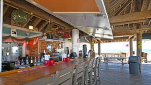 Eine Strandbar mit Holzbalken, einer Theke mit verschiedenen Flaschen und Blick auf das Meer.