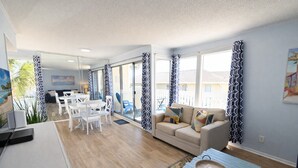 Ein modernes Wohnzimmer mit Sofa, Couchtisch und Essbereich. Ein Balkon mit gemusterten Vorhängen und Blick ins Freie.