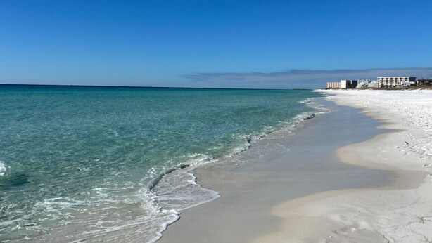 Ein Strand mit klarem, blauem Wasser, Sandküste und einem Gebäude im Hintergrund.