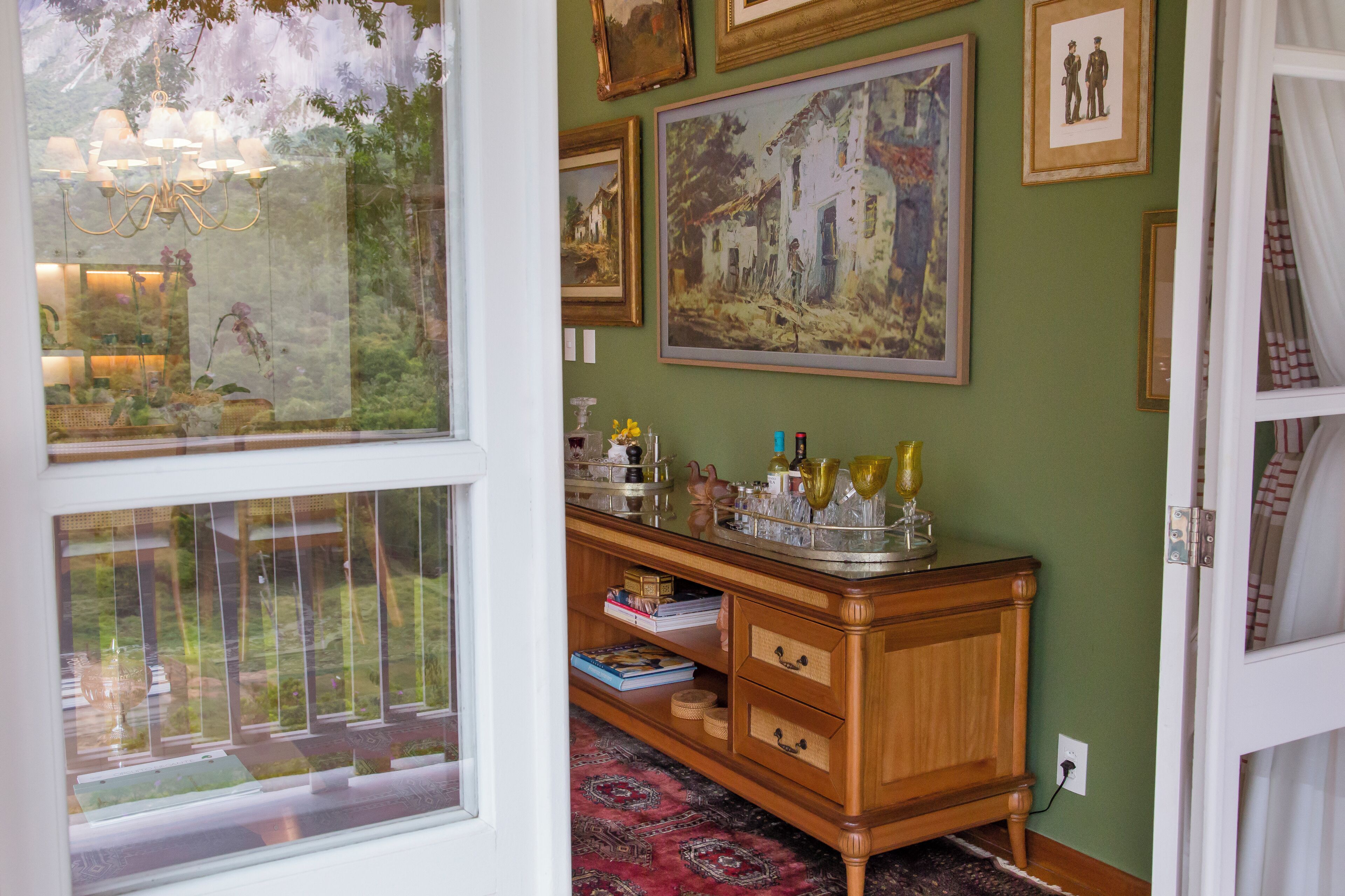 Una habitación con un ventanal grande, un mueble de madera con cristalería y cuadros en la pared.