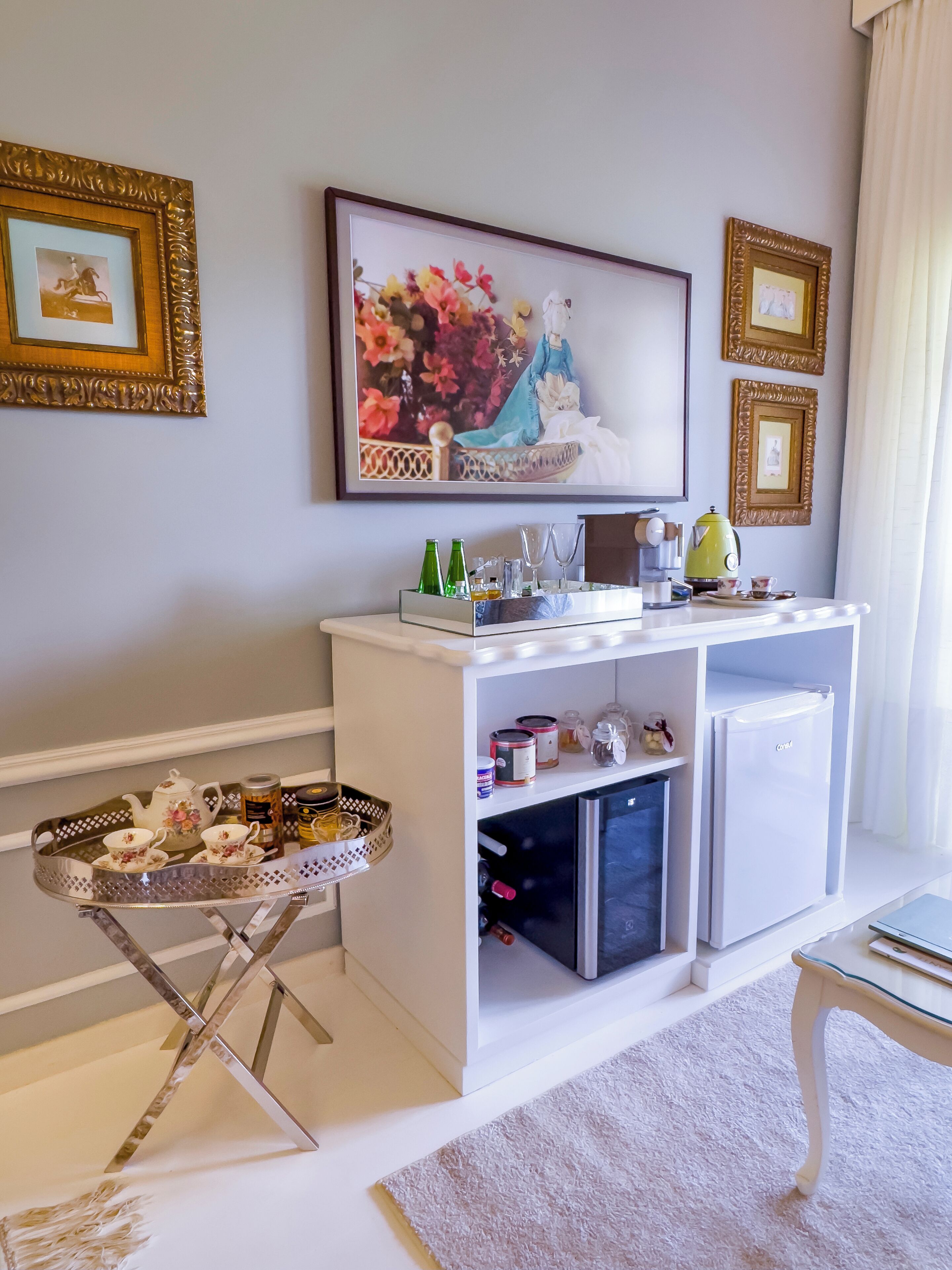 A room with a white console table, a small refrigerator, a glass tray with tea set, and framed artwork on the wall.