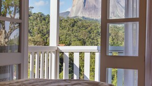 A room with a bed, a balcony with a view of a mountain and forest.