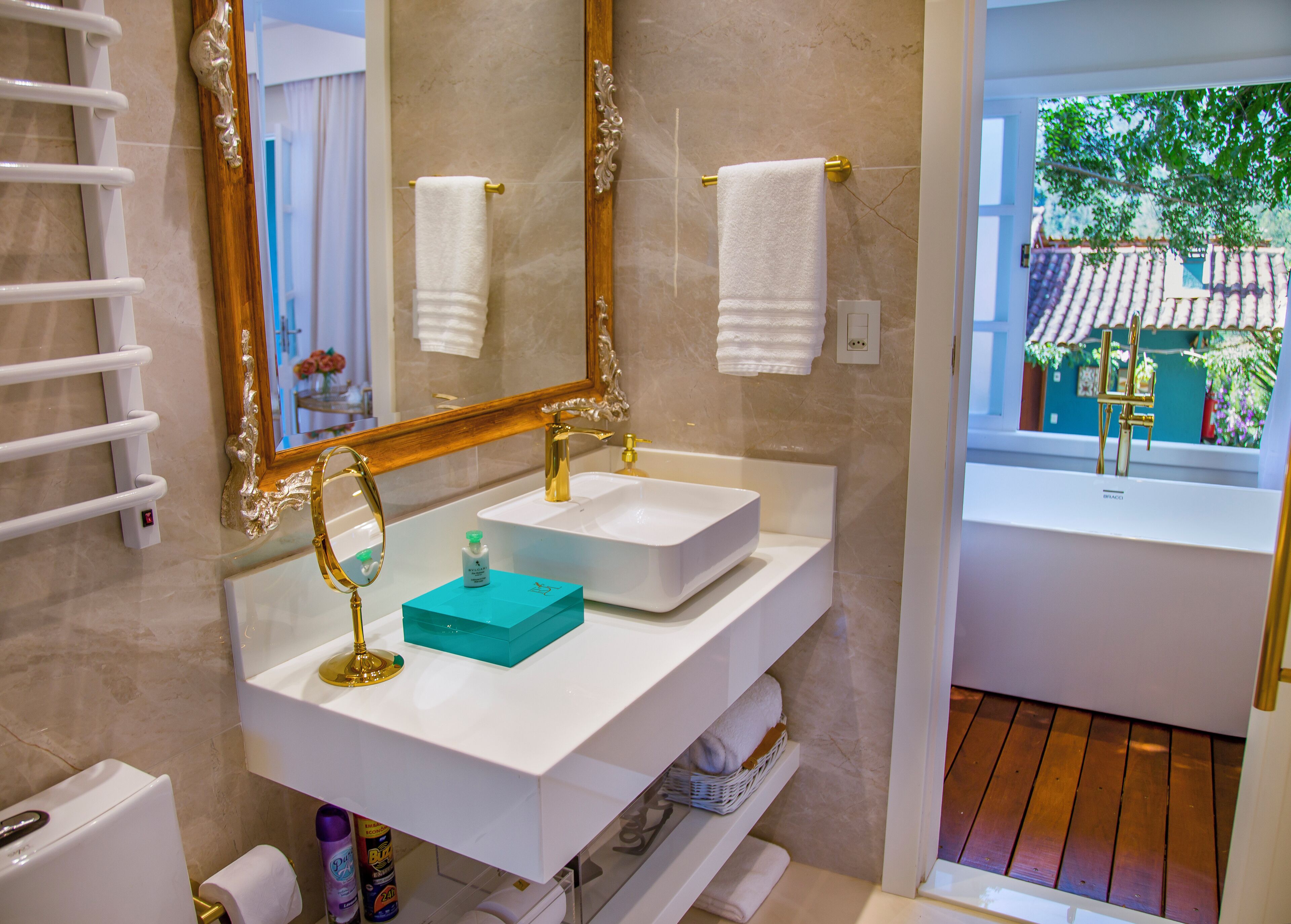 Un baño con lavamanos blanco, detalles dorados, un espejo y vistas a un patio empedrado.