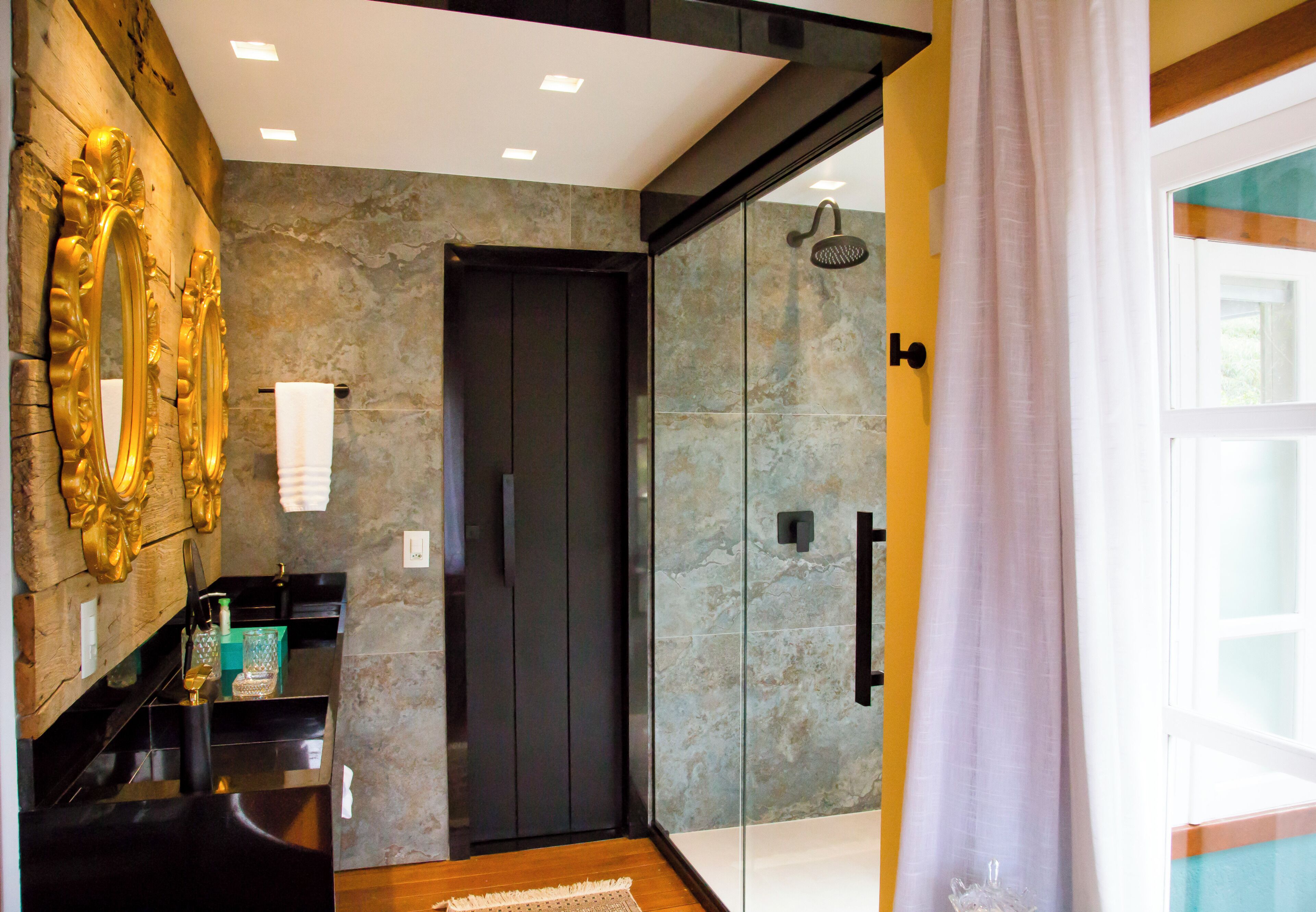 Un baño moderno con cabina de ducha de vidrio, puerta oscura y espejo con marco dorado.