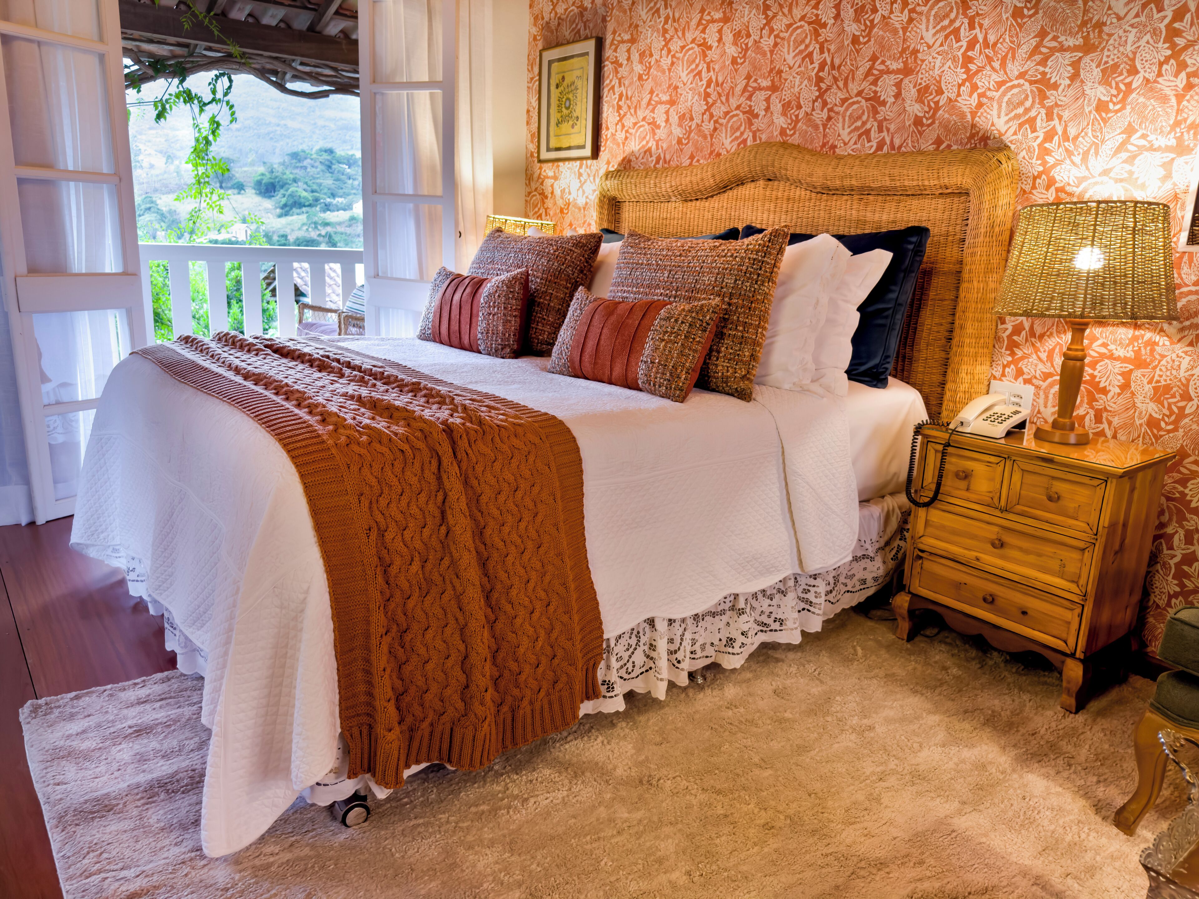 A bedroom with a large bed, a nightstand with a lamp, and a view of greenery outside.
