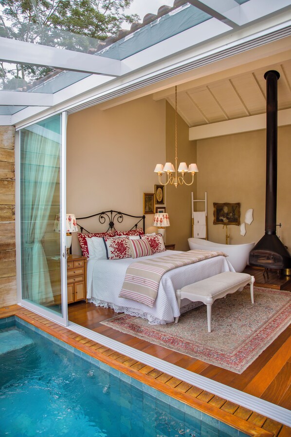 A bedroom with a glass door leading to a pool, a bed with white bedding, a chandelier, and a fireplace. - Pousada Rabo do Lagarto (Domingos Martins)