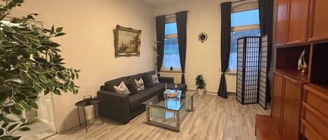 Uma sala de estar com sofá, mesa de centro de vidro, planta e um quadro na parede.