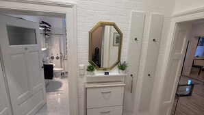 Un couloir avec un mur en briques blanches, un miroir, une commode blanche avec des tiroirs et une porte donnant sur une autre pièce.