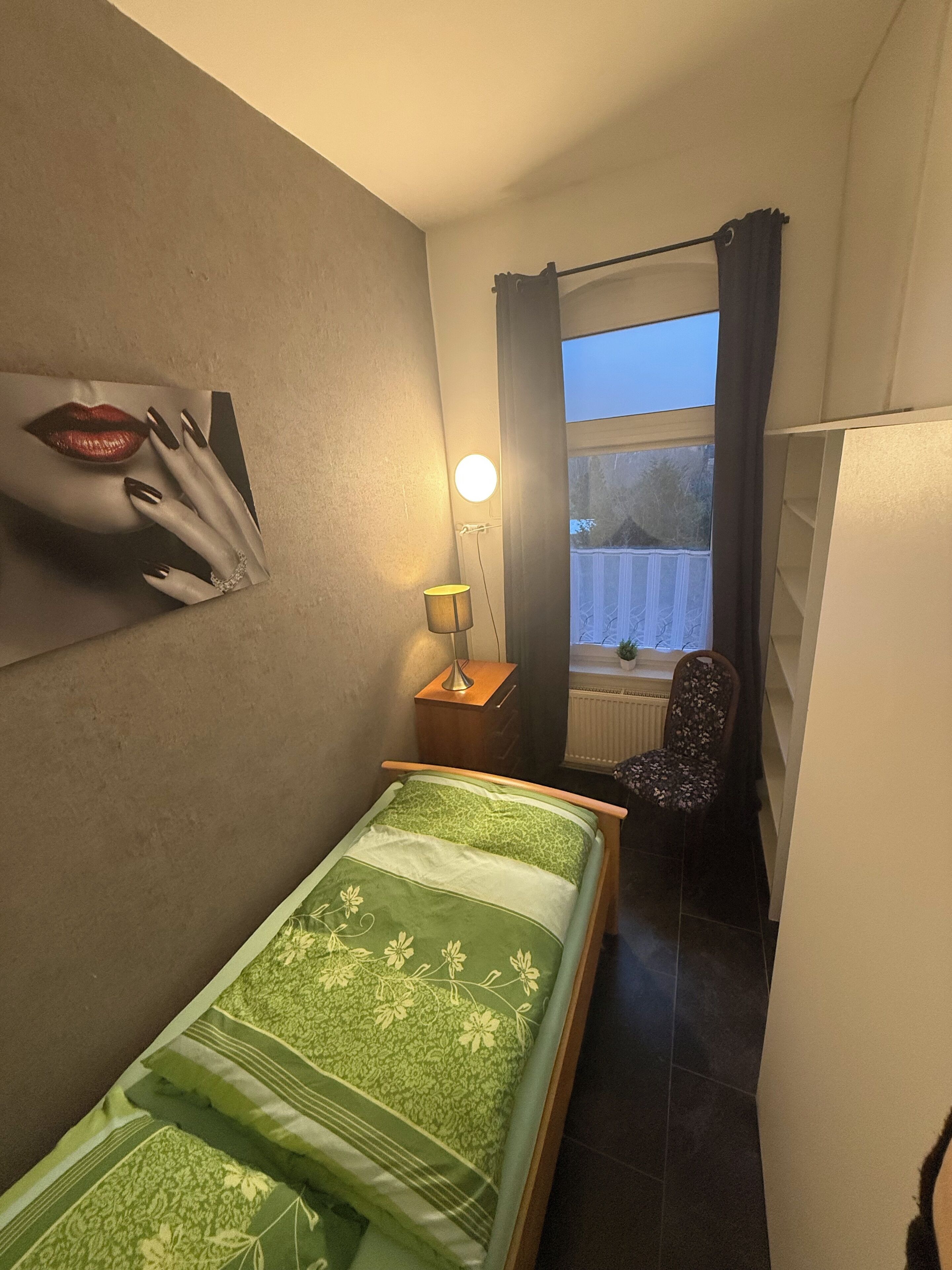 A small, neatly arranged bedroom with a bed, a chair, a desk, and a lamp.