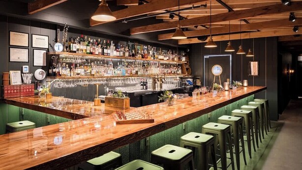 Un'area bar con un bancone in legno, sgabelli verdi e una selezione di bottiglie e bicchieri.