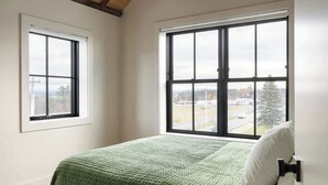Una camera da letto con soffitto in legno, un letto grande con copripiumino verde, due finestre e vista sul paesaggio.