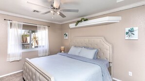 A bedroom with a bed, a ceiling fan, a window with sheer curtains, and a wall-mounted shelf with a plant. - Havasu Tropics - Pool, Spa, Game Room, Sleeps14 (Lake Havasu City)